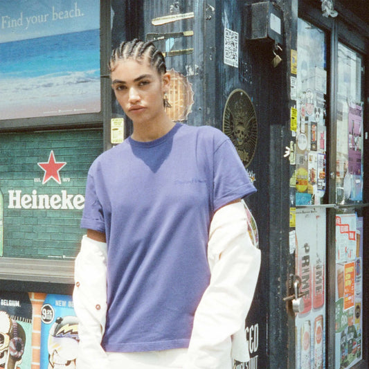 Person wearing a purple shirt with a white long-sleeve underneath, standing in front of a store with various signs and advertisements.