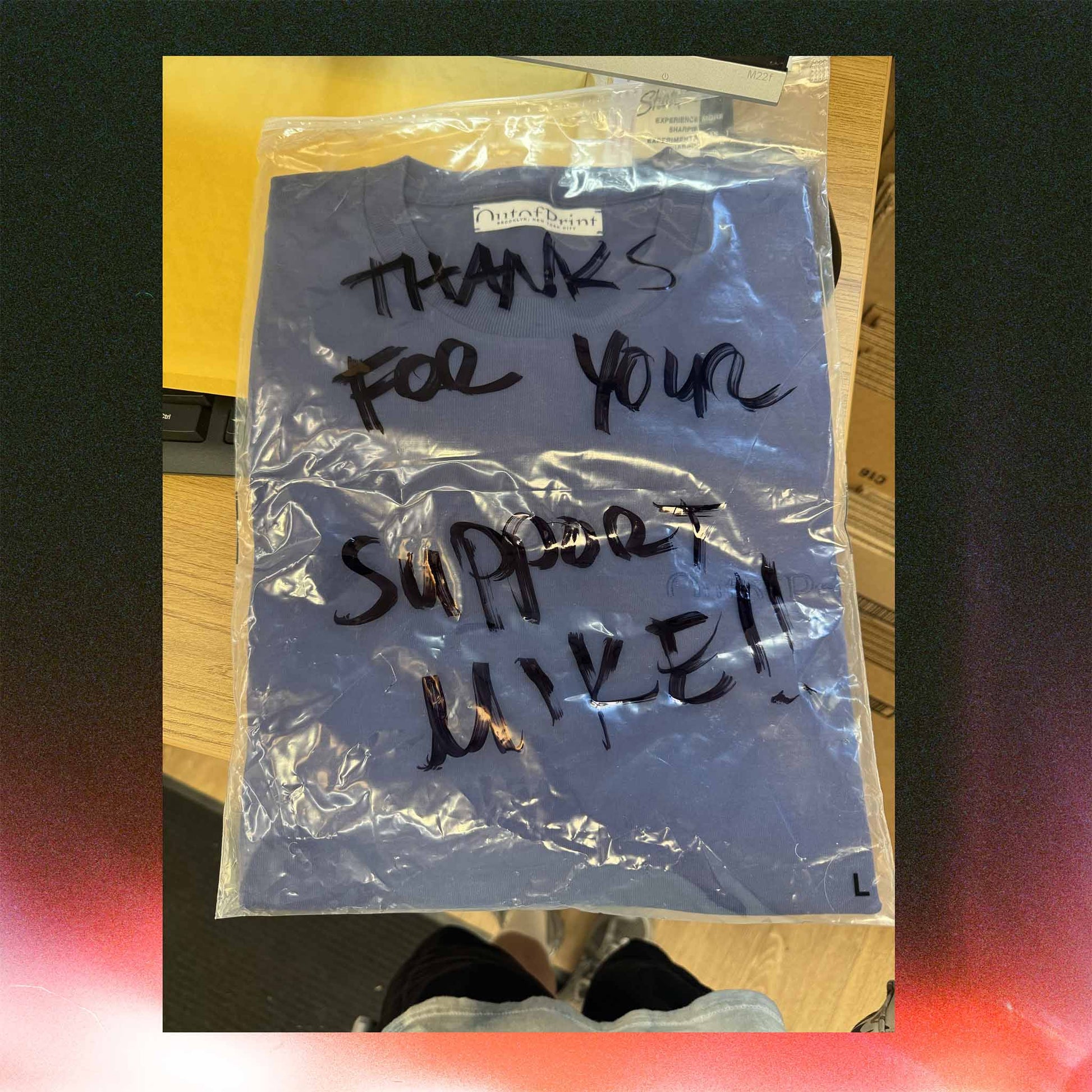Blue t-shirt in a clear plastic bag with handwritten text on a wooden surface.