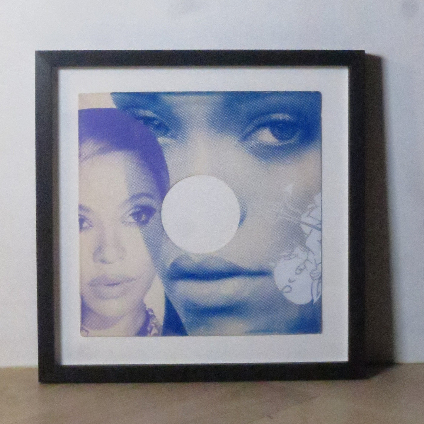 A square black-framed picture frame with a white mat, containing a blue-toned collage print, photographed leaning against a light wall on a wood floor.