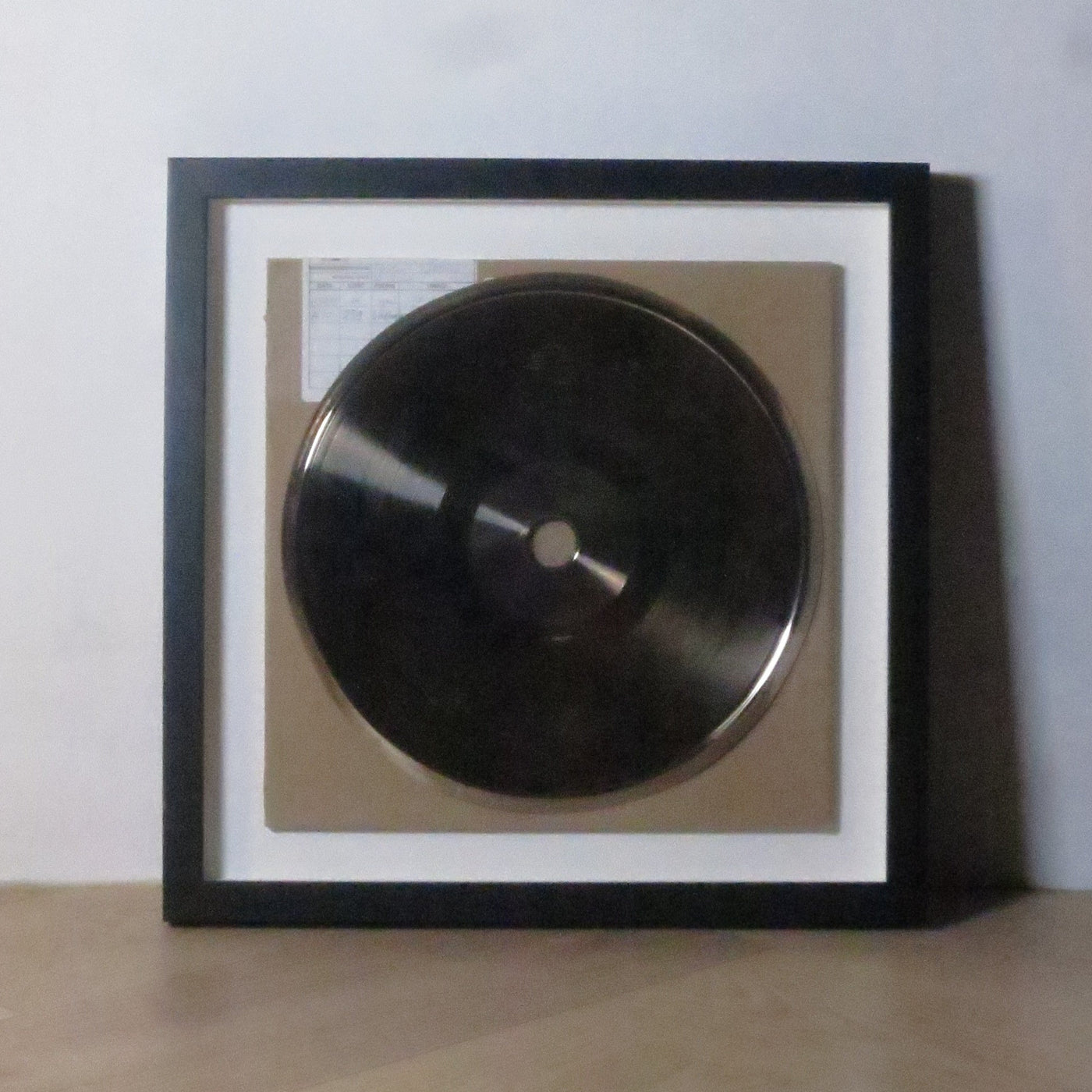 A framed silver vinyl plate display mounted on a light gray wall, with a light wood floor visible on the right side.
