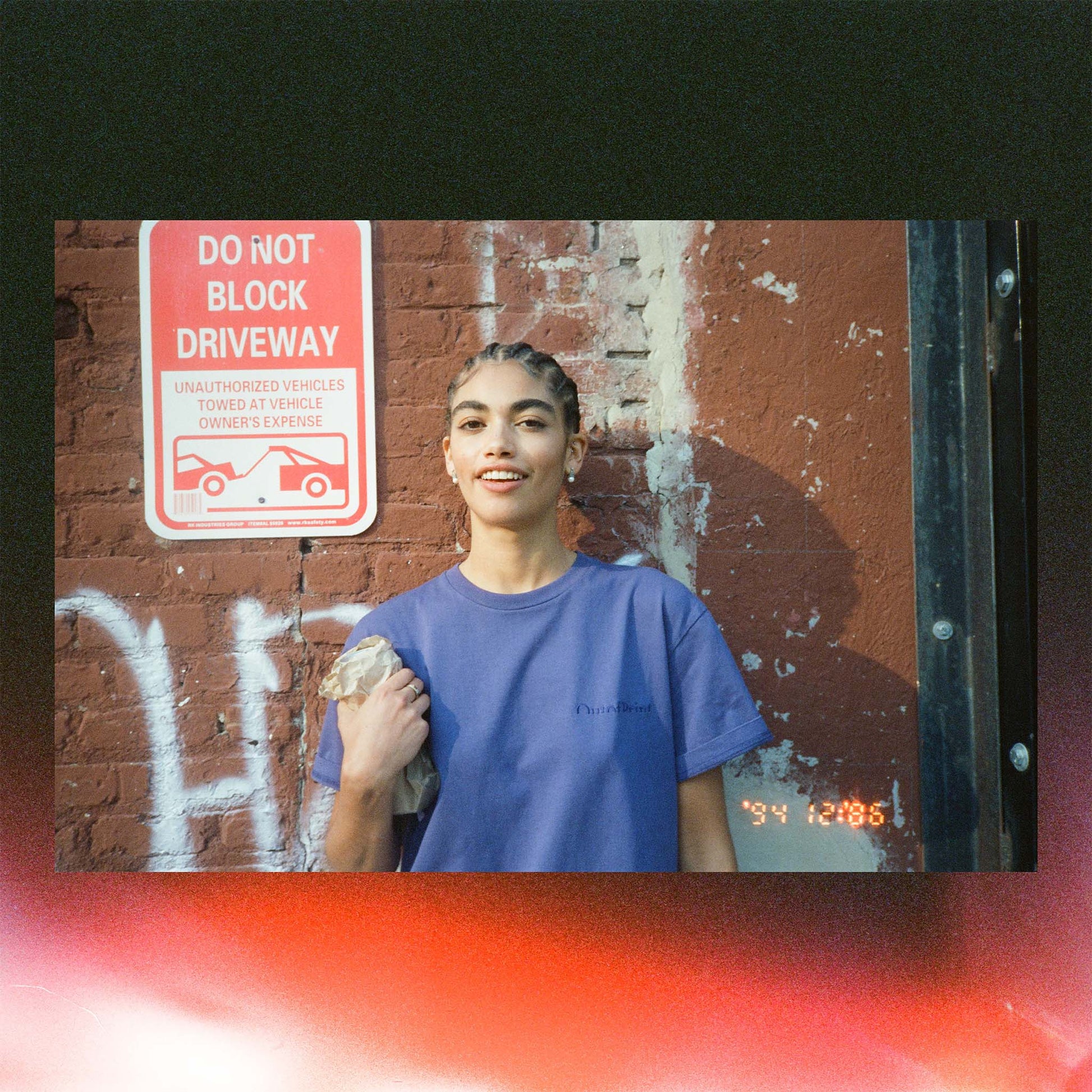 Person wearing a purple shirt standing in front of a red 'Do Not Block Driveway' sign on a brick wall.