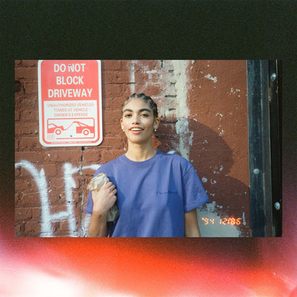 Person wearing a purple shirt standing in front of a red 'Do Not Block Driveway' sign on a brick wall.